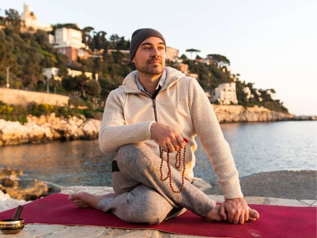 Homme méditant en position du lotus sur un tapis de yoga, tenant un mala, près de la mer au coucher du soleil, avec des maisons en arrière-plan.