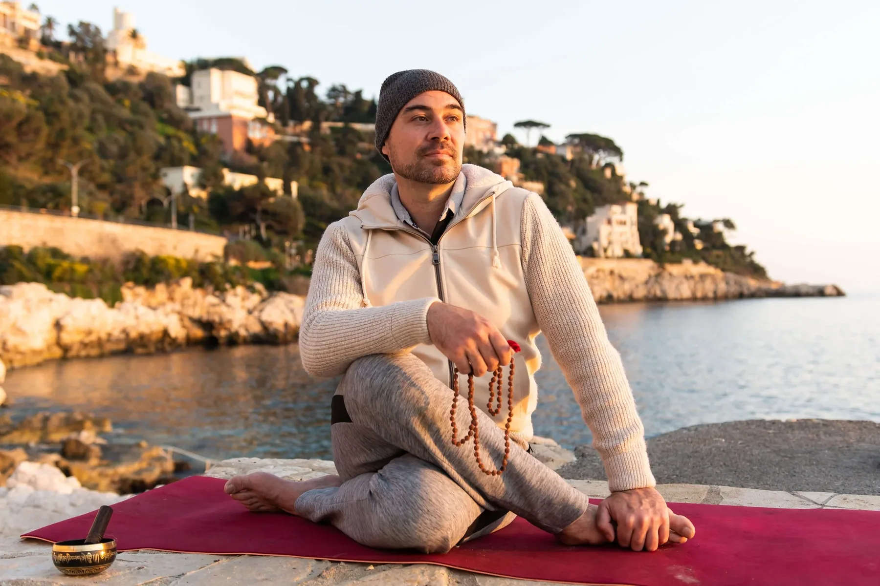 Un homme assis en position de méditation sur un tapis rouge près de la mer au coucher du soleil, tenant un mala en bois. Il porte un sweat à capuche beige, un bonnet gris, et des vêtements de sport gris. Un bol de méditation est placé à côté de lui.