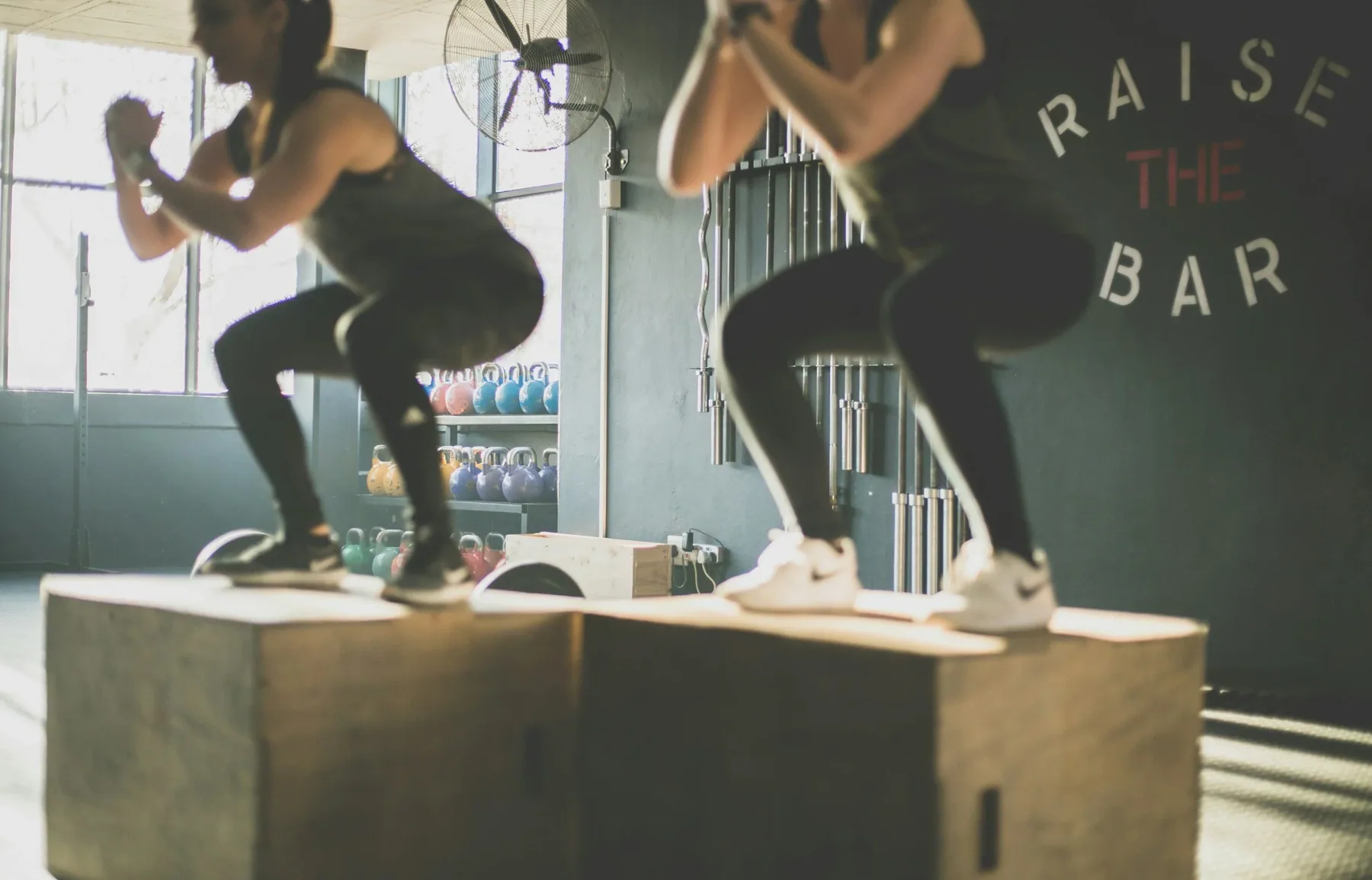 Deux femmes en tenue de sport effectuant des squats sur une plateforme dans une salle de sport, avec des kettlebells colorés en arrière-plan et un mur avec le logo 'Rai­se The Bar'.